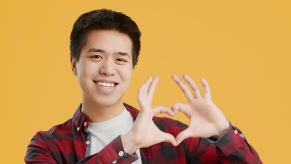 Cheerful Asian Guy Showing HeartShape Gesture Posing Over Yellow Background alt