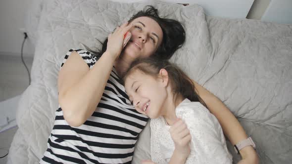 The Daughter Hugs Her Mother Lying Together on the Bed and Communicating on a Mobile Phone with