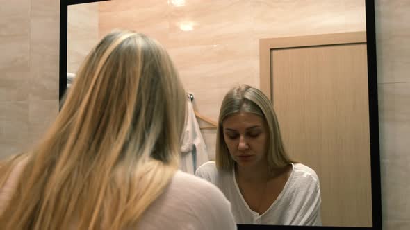Tired Young Blonde Woman Looks at the Reflection in the Mirror and Washes Her Face alt
