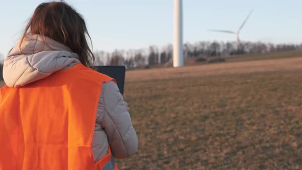 Female Engineer in Orange Vesta Goes to Wind Turbines with a Tablet to Checks Their Operation alt