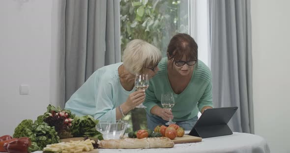 Two Cheerful Smiling Senior Women with Wine Glasses Looking at Tablet Screen and Talking alt