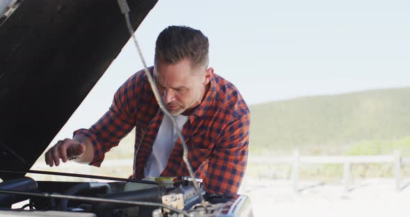 Stressed caucasian man looking at broken down car with open bonnet on sunny day at the beach alt