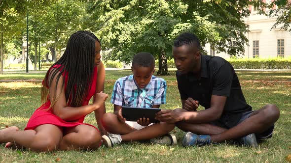 A Young Black Family Sits on Grass in a Park and Works on a Tablet alt
