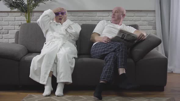 Mature Blond Caucasian Woman in White Bathrobe Sitting on Sofa and Emotionally Watching 3D TV alt