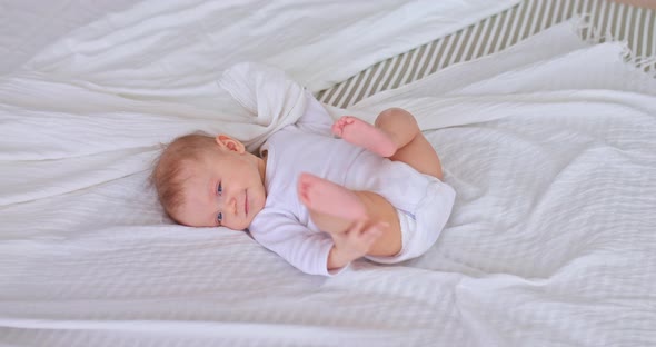 Pretty Baby is Playing with Arms and Legs Lying in a Bodysuit on a White Bedspread alt