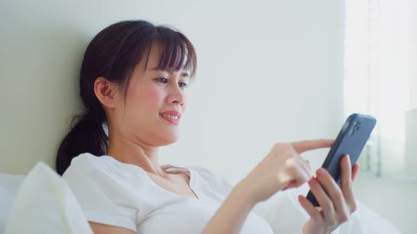 Asian beautiful woman laying on bed, using mobile phone-click and swipe on screen in house. alt