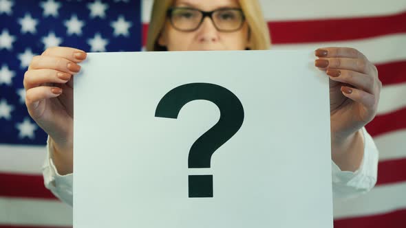 A Woman Holds a Poster with a Question Mark Against the Background of the American Flag alt