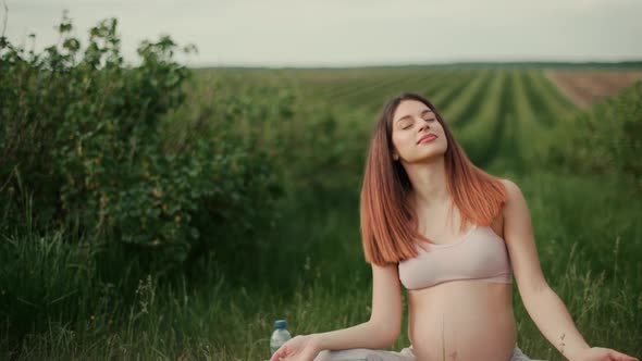 Pregnant young woman with dark hair in a pink top and light pants enjoys the sun outdoors. 