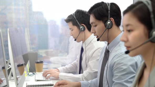 Young Asian male telemarketing operator with team working at call center office in the city