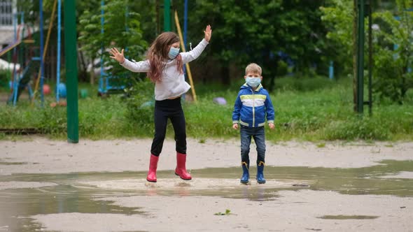 Little girl and a boy in medical masks and rubber boots jump through puddles on a spring day