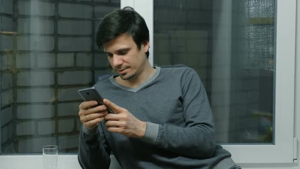 Man Is Browsing Internet Pages on Mobile Phone Standing Near the Window on the Balcony.
