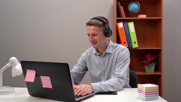 A Young Worker Communicates with a Client at an Online Conference From the Office Through Headphones alt