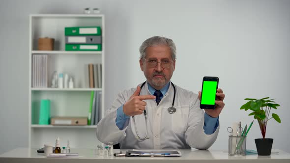 Aged Male Doctor Holds Smartphone with Green Screen Chroma Key Points at It with Finger alt