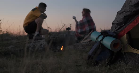 Unfocused Two Young Travelers Man and Woman Sitting Near the Campfire and Tent Outdoors During