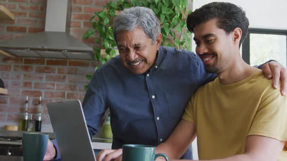 Smiling biracial man with arm around young son using laptop at home alt