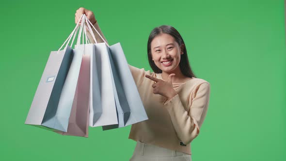 Asian Woman Holding Shopping Bags Up Before Pointing And Smiling At Green Screen Studio alt