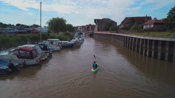 Man kayaking on meandering river next to boats and houses, drone closely follows and pans up to reve alt