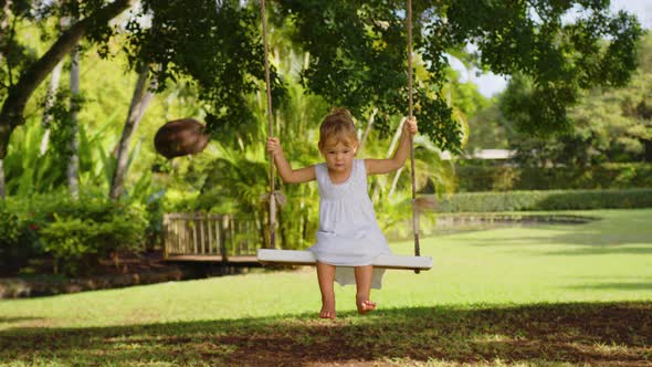 A Little Girl in a White Dress Rides on a Rope Swing in the Park alt