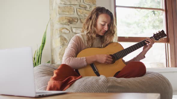 Happy caucasian woman playing acoustic guitar, sitting on beanbag in sunny cottage living room alt