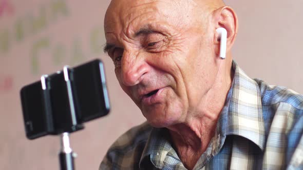 portrait of a caucasian pensioner 70-79 years old in airpods holding a smartphone and emotionally ch alt