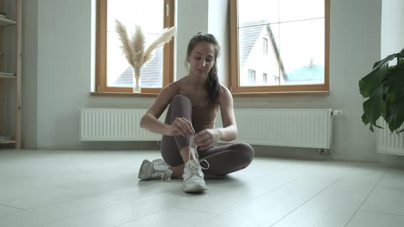 Young Girl in Sportswear Sitting on Floor on Knees Tying Shoelaces on Sneakers