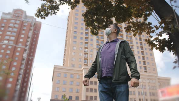 A Man in a Medical Mask Looks Up Against the Backdrop of Downtown Highrise Buildings