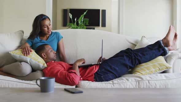 Happy biracial couple on sofa with laptop and talking alt