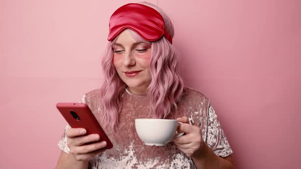 Portrait of Joyful Sleepy Woman Wears Red Eye Mask and Pink Pajamas Uses Cell Phone Connected to