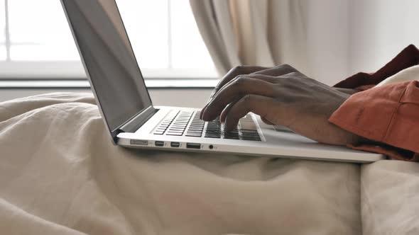 AfricanAmerican Man Types on Laptop Keyboard Sitting on Bed alt