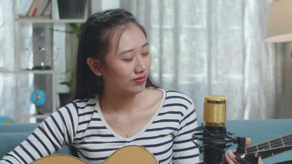 Close Up Of Asian Woman With Microphone Having Video Call On Laptop While Playing A Guitar At Home alt