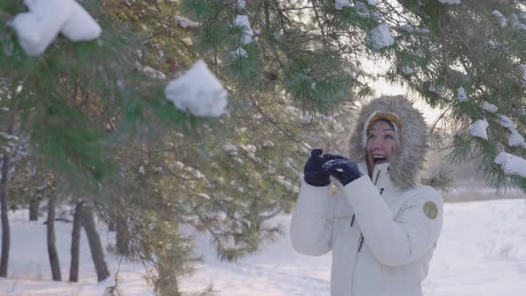 Cute Girl in Winter Down Coat Crushing Snow From a Pine Branch and Rejoices alt