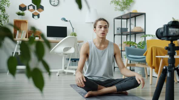 Male Yoga Teacher Vlogger Recording Tutorial Using Camera in Modern Apartment Talking Sitting on Mat alt