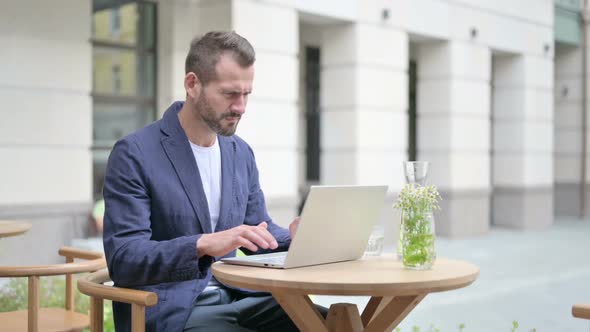 Man Reacting to Loss While Using Laptop Sitting in Outdoor Cafe alt