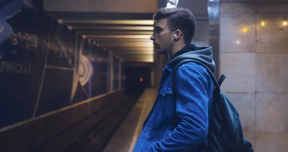 Young Man in a Jeans Jacket and Glasses is Waiting for a Train at the Metro Station alt