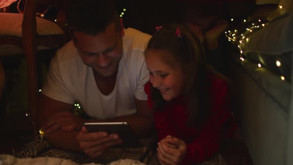 Caucasian father and daughter lying and using digital tablet under blanket fort during christmas alt