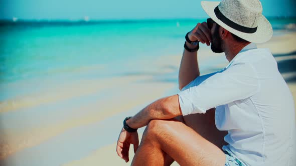 Man Sitting On Tropical Beach In White Shirt. Handsome Man Relaxing Lounging On Caribbean Beach. alt