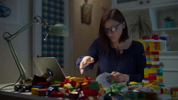 Young 30s working from home mother with pile of kids toys on the working table with laptop  alt