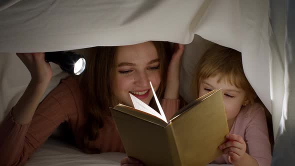 Young Mother Reading Goodnight Story Fairytale to Child Daughter Under Duvet Blanket in Night Room alt