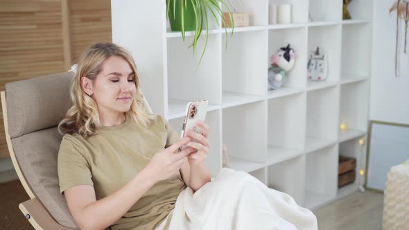 Cute young blonde woman uses phone while sitting at home in armchair Using technology concept alt
