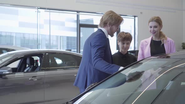 Confident Salesman in Suit and Two Female Clients Talking Near Modern Car. Women Choosing Vehicle alt