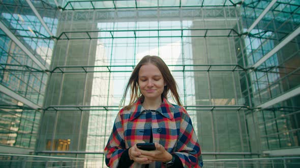 Tired Young Woman Walks in Modern Glass Building Using Smartphone App