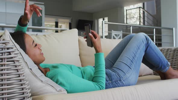 Mixed race woman lying on couch having video chat on smartphone blowing kisses alt