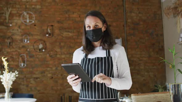 Caucasian female business owner wearing face mask and apron, using tablet, looking at camera alt