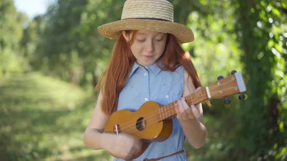 Medium Shot Portrait of Happy Skilled Caucasian Redhead Girl Singing Playing Ukulele Outdoors alt