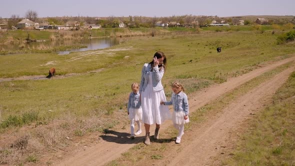 Mother and Her Twin Daughters Walk Along a Country Road on a Farm in the Spring. alt