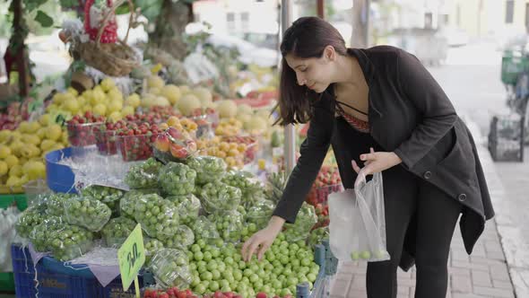 Young attractive woman shopping at the greengrocer choosing plum alt