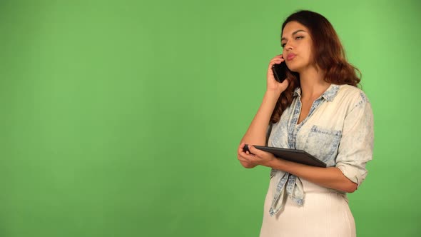 A Young Beautiful Caucasian Woman Talks on a Smartphone and Holds a Tablet  Green Screen Background