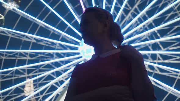 Woman Looks Thoughtfully at the Camera on the Background of the Ferris Wheel