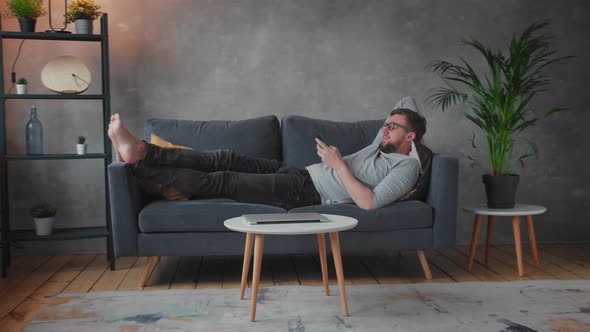 Young Man with Glasses is Lying on the Couch and Texting on the Phone alt