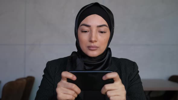 Joyful Young Muslim Woman in Hijab is Playing Video Game in Smartphone